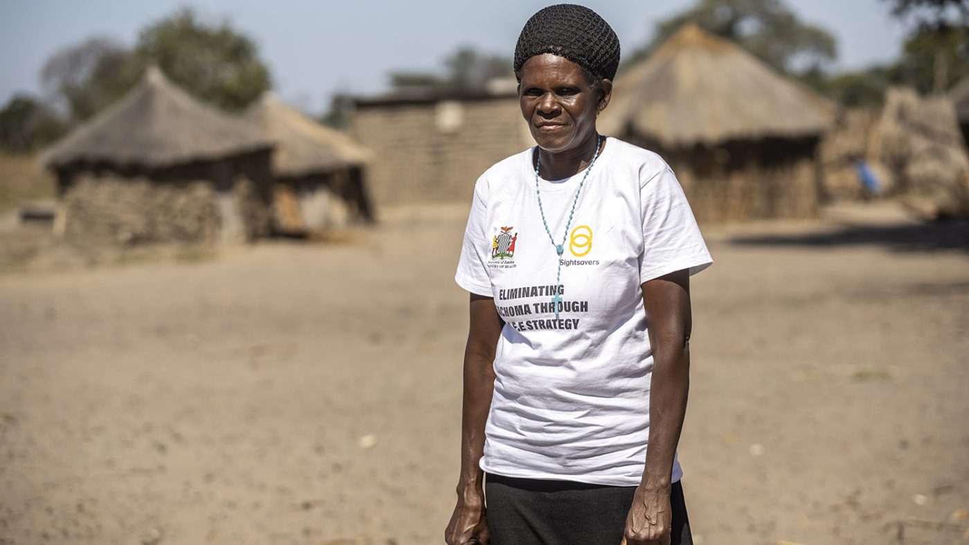 Zita, a community health worker, stands outside in a village. There are grass hut homes behind her.