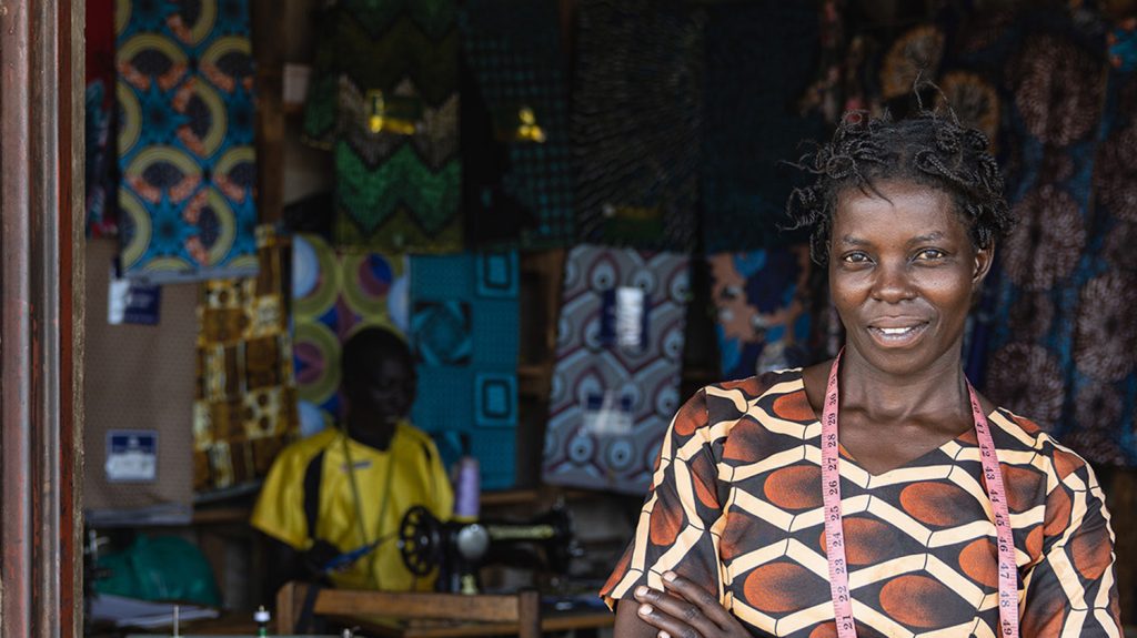 A woman with a tape measure around her shoulders stands outside a workshop.