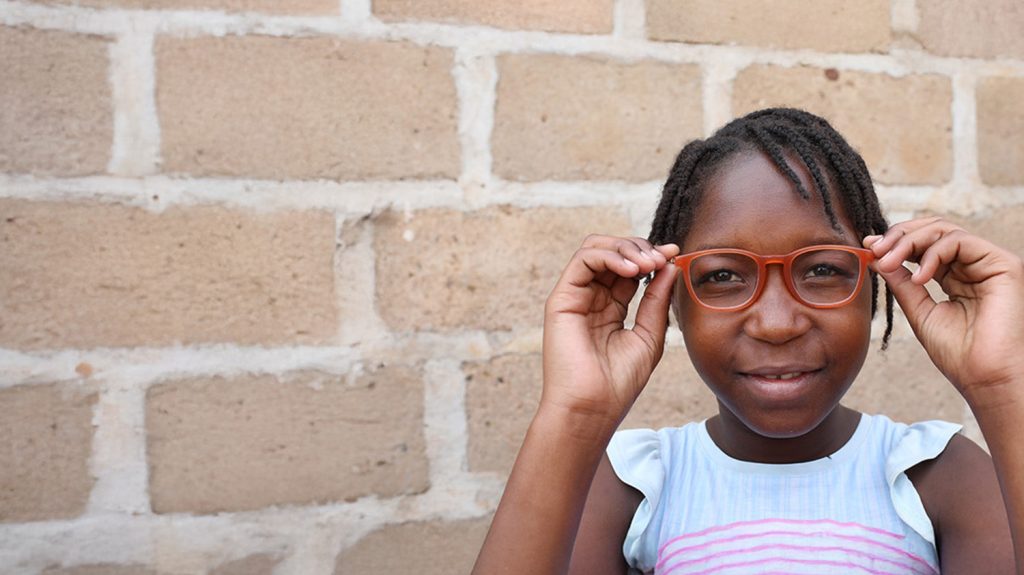 A girl wearing red glasses stands in front of a brick wall.