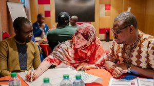 A woman wearing a hijab writes on flipchart paper during a workshop. Two men are sitting on either side of her, watching on as she writes.