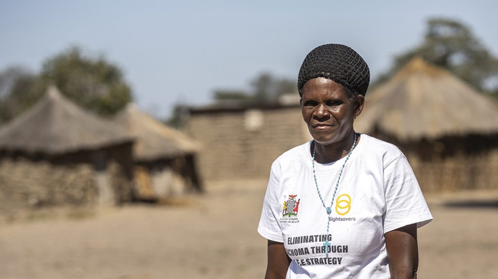 A women wearing a Sightsavers T-shirt stands outside. Behind her are traditional grass homes.