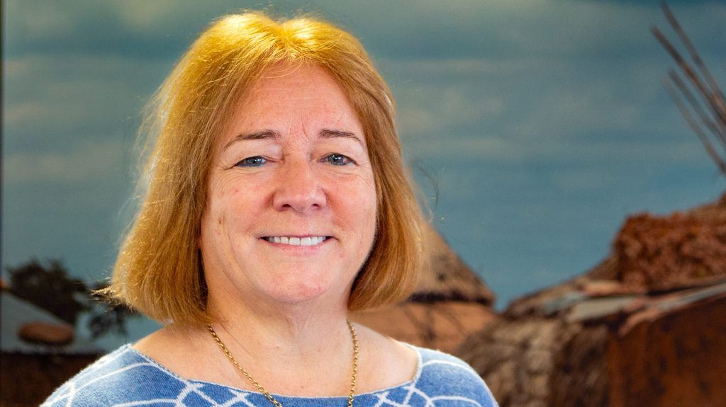 Sightsavers CEO Caroline Harper stands in front of background photo depicting a traditional village with grass huts.