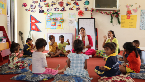 In a classroom, a teacher speaks to a group of young children sitting in a circle.