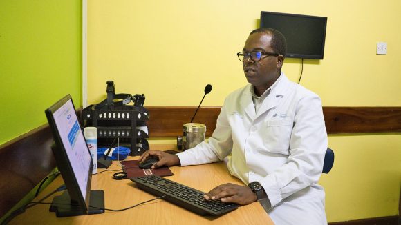 A man wearing a white medical coat sits in front of a computer at a desk.