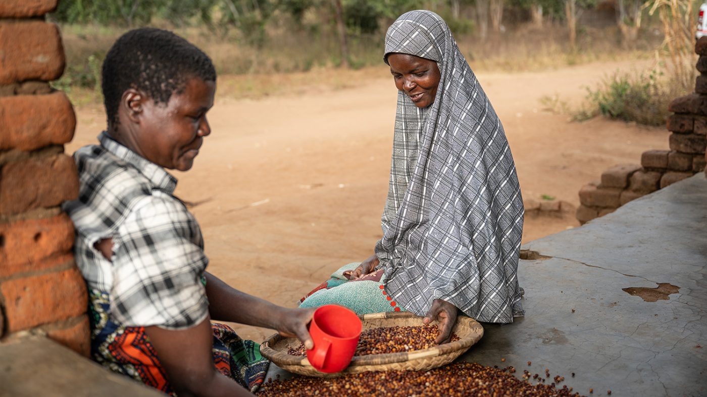 Ruth sits on a porch sorting through grain in a wicker basket. A man is measuring the grain using a plastic cup.