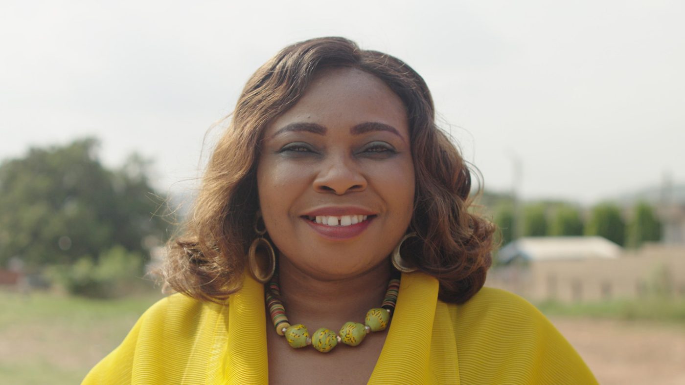 Veronica smiles broadly. She is wearing a bright yellow jacket and a beaded necklace.