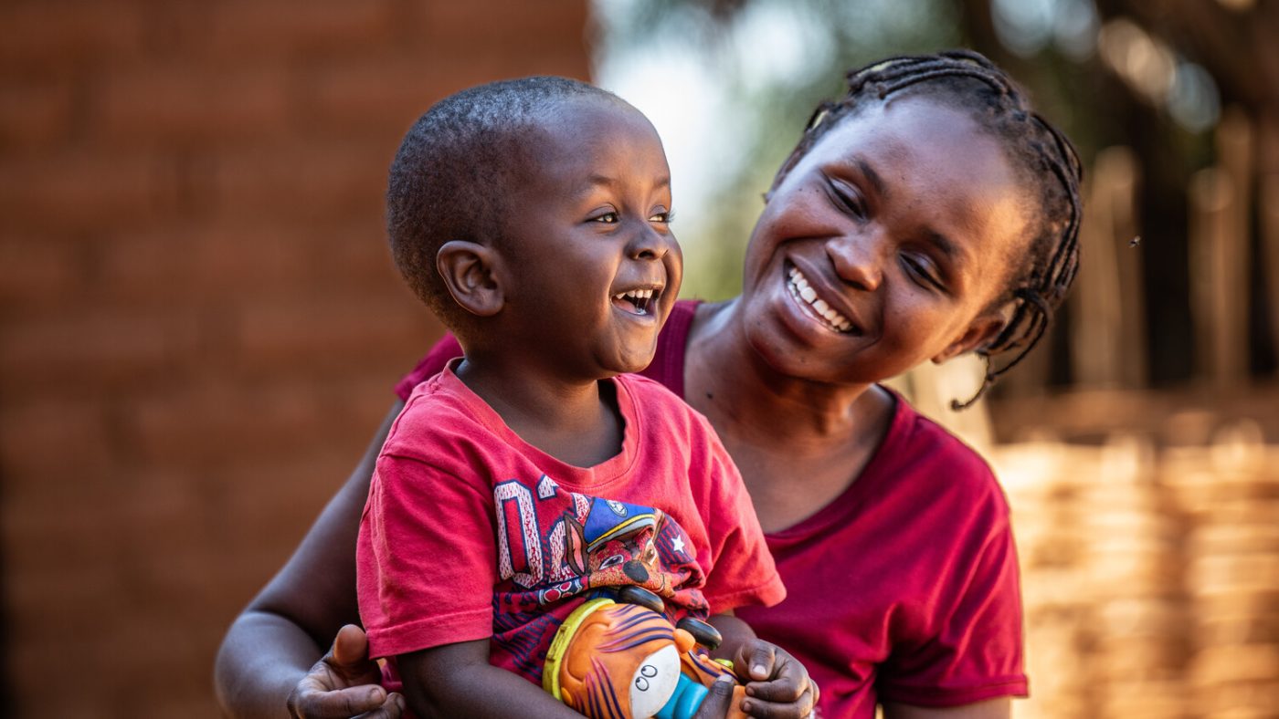 Three-year-old Omega sits with his mum, Jane, holding a toy. They are both laughing together.