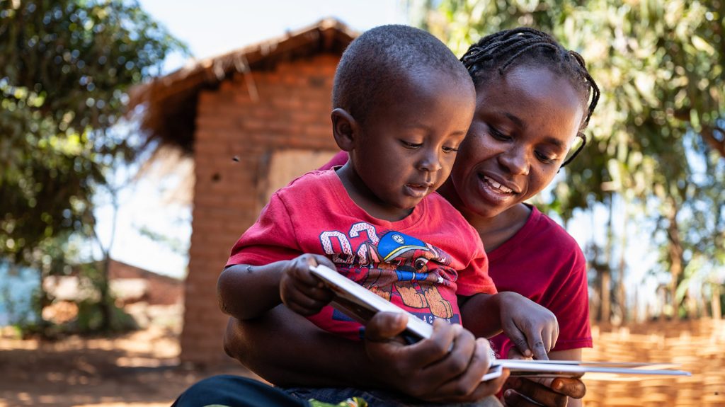 Omega sits on his mum Jane's lap. They are reading a book together, pointing to the pictures.