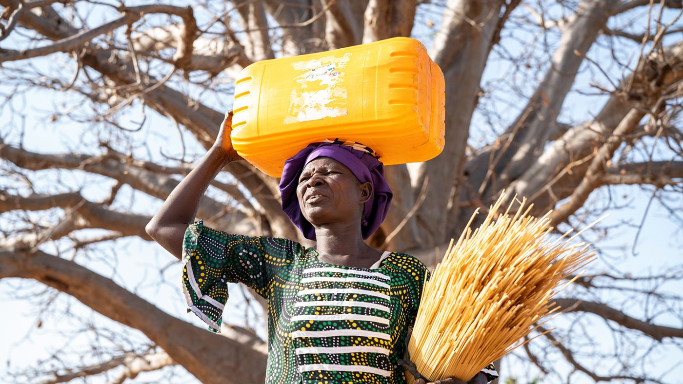 Mabila stands under a tree with a sheaf of reeds in one arm. She is carrying a large plastic container on her head.