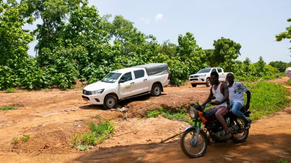 Two men ride on a motorbike along a dusty track. Two 4x4 vehicles drive alongside them.
